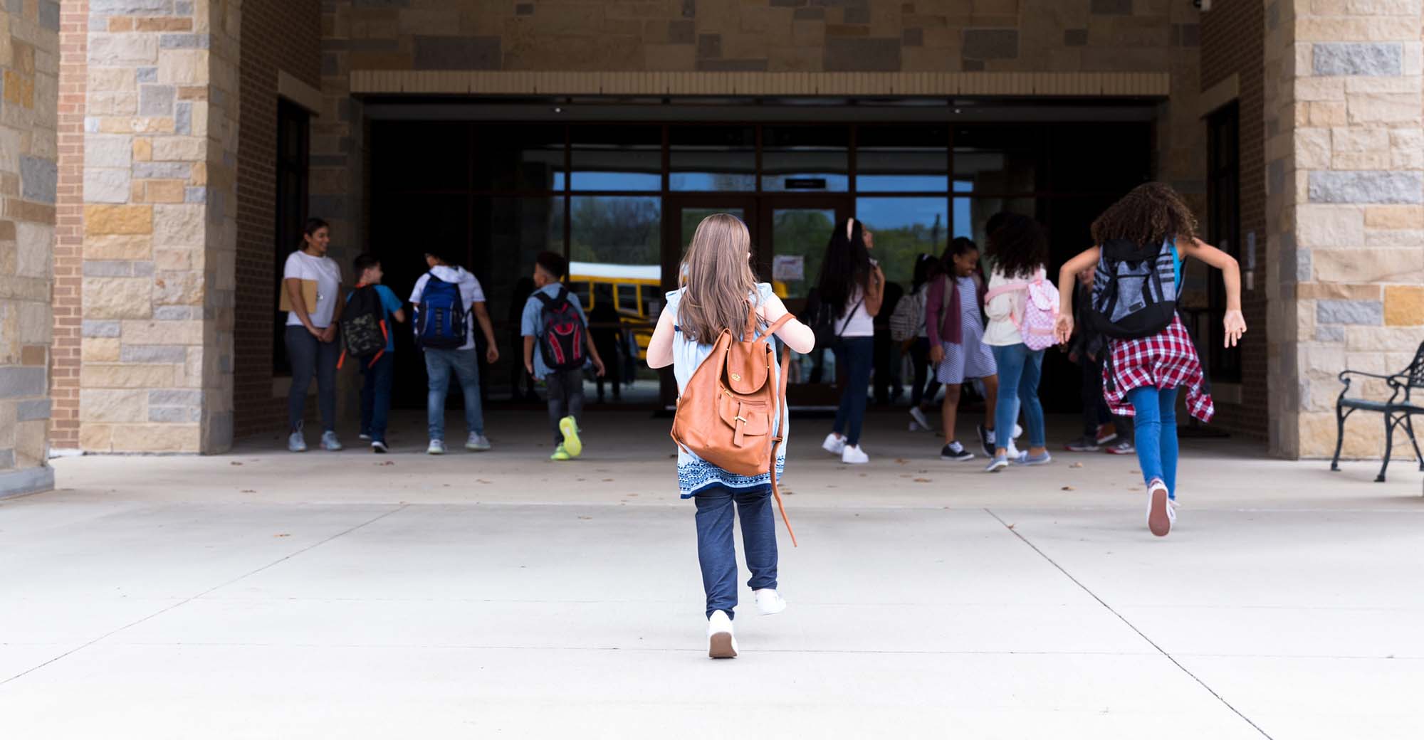 Rear view of elementary school girl walking into the school building. Other students walk into the building ahead of her.