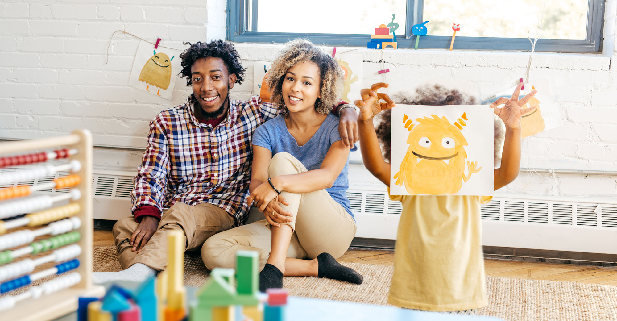 A Black family is sitting in their child's playroom. The parents are sitting along the back wall while their child stands in the foreground and holds up a painting of a yellow, smiling monster.