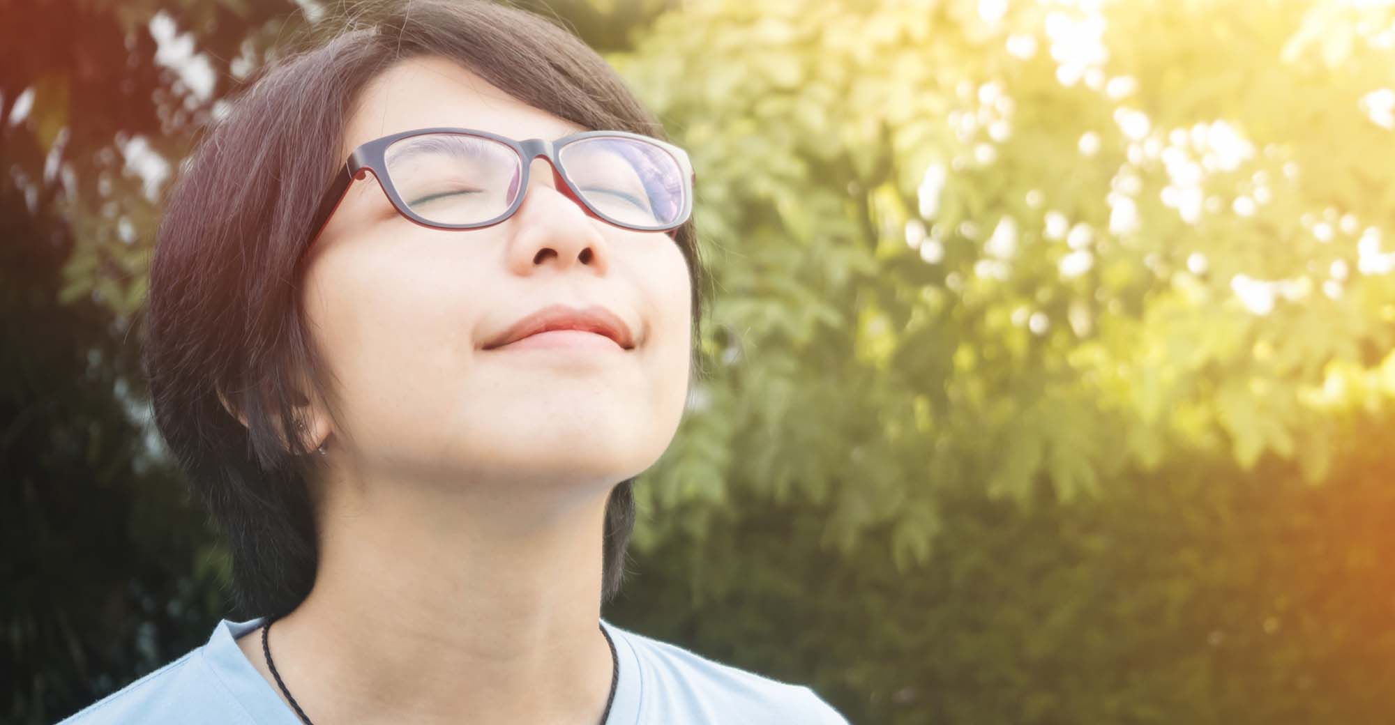 A person wearing glasses looks up toward the sunlight while smiling.