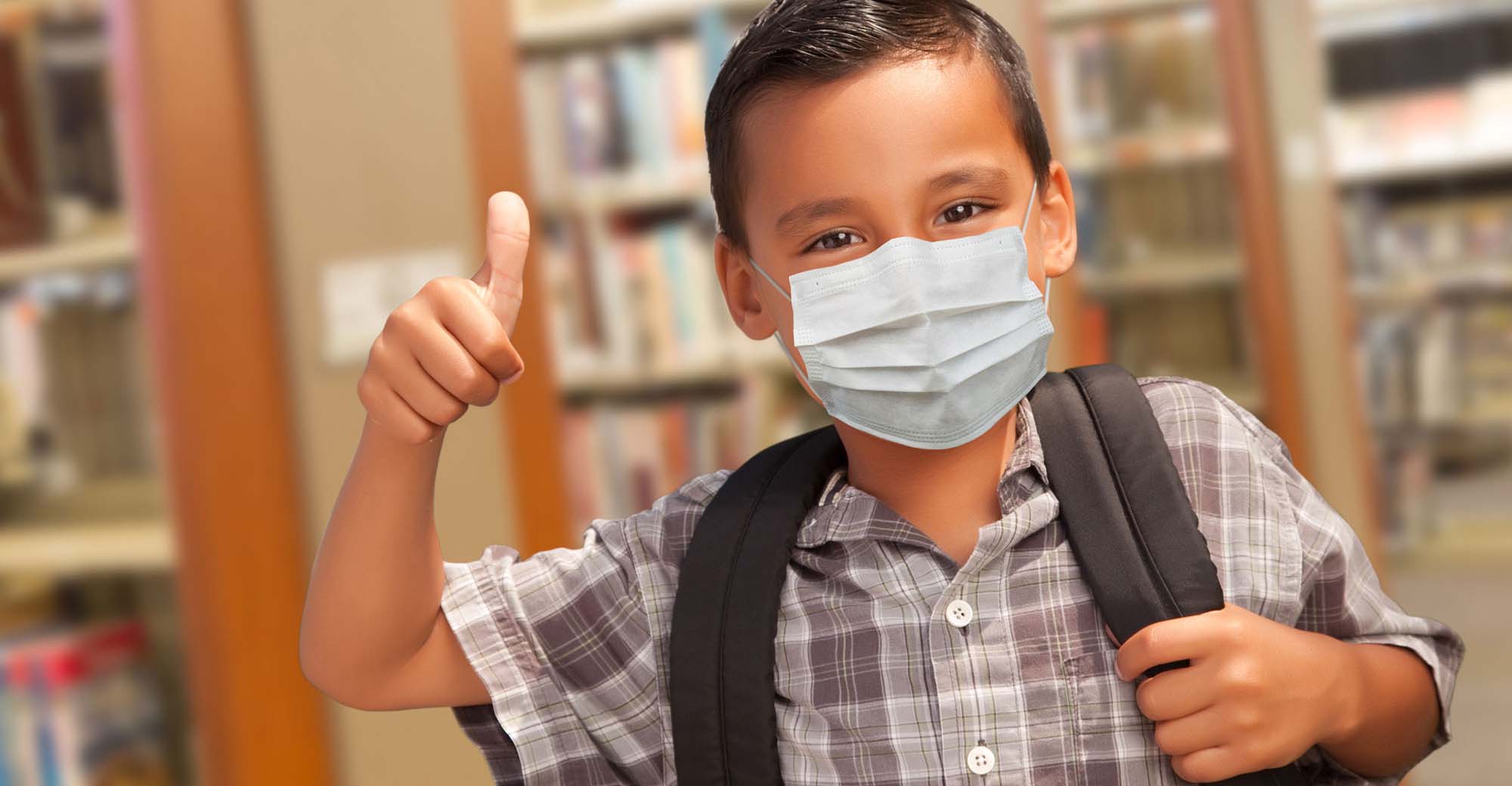 A Hispanic boy wears a medical face mask and looks toward the camera holding his thumb up.