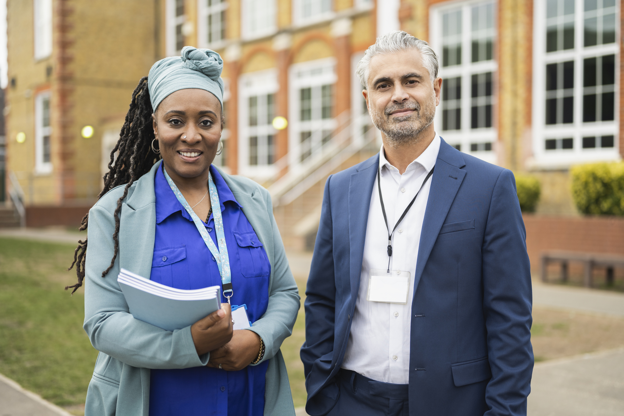 Waist-up front view of mature Black female and multiracial male colleagues standing side by side in front of school building and smiling at camera.