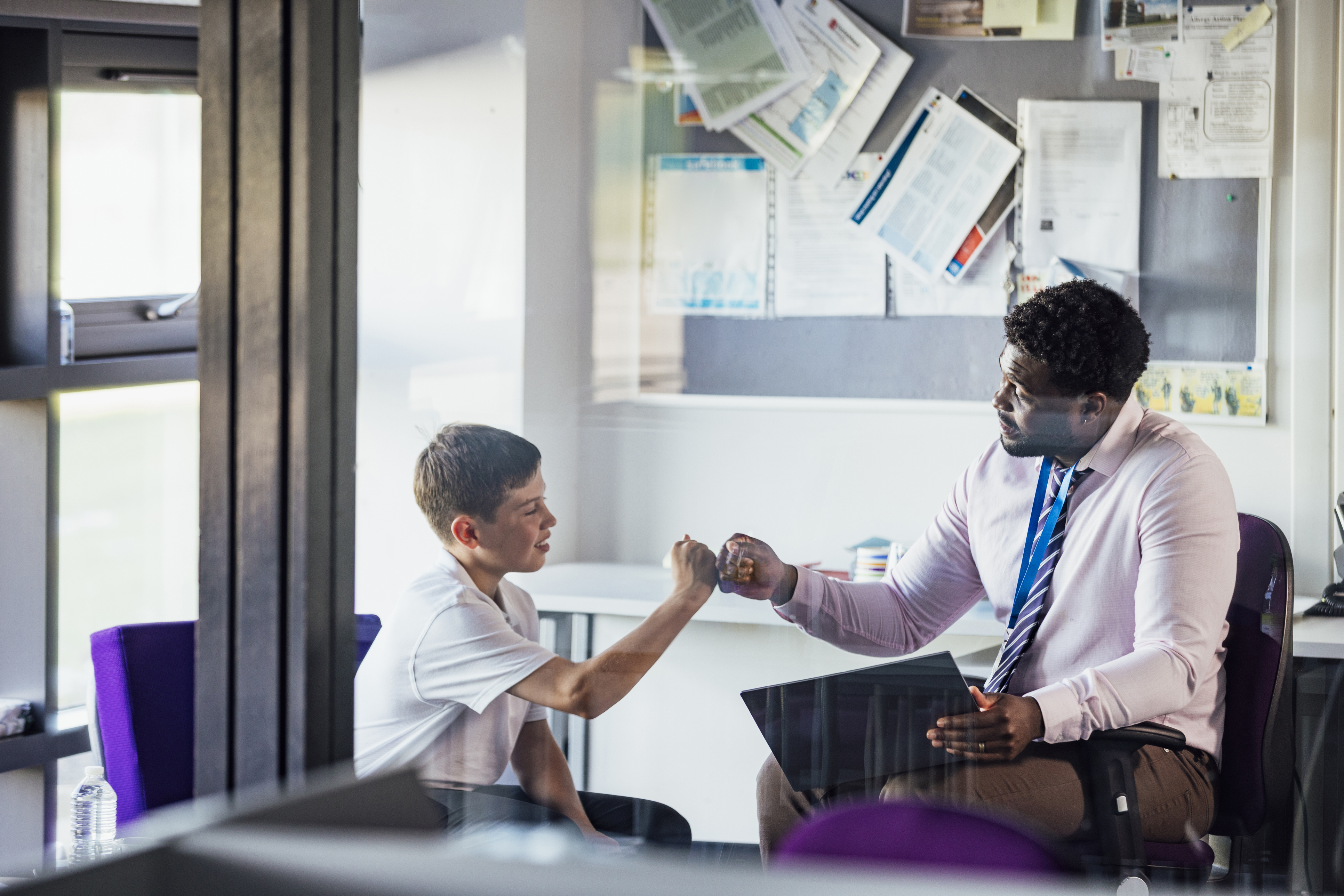 A male secondary school student sitting in an office in the school, having a meeting with his teacher. They are giving each other a fist bump after a successful meeting.